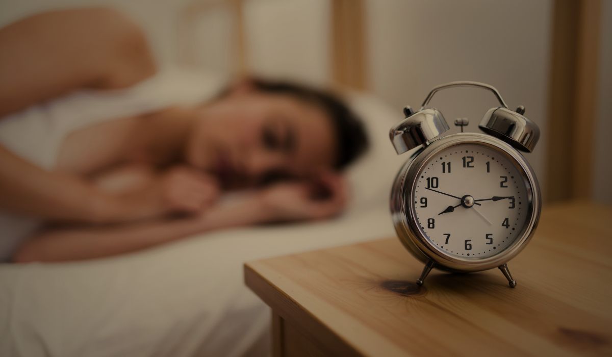 Relógio despertador ao lado da cama com pessoa dormindo ao fundo, ilustrando higiene do sono o que e e hábitos para dormir melhor