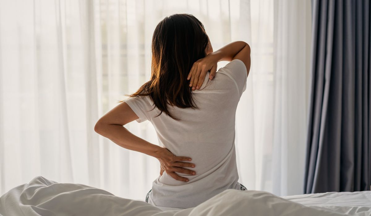 Pessoa sentada na cama com a mão nas costas e no ombro, ilustrando a busca pelo melhor colchão para dor nas costas
