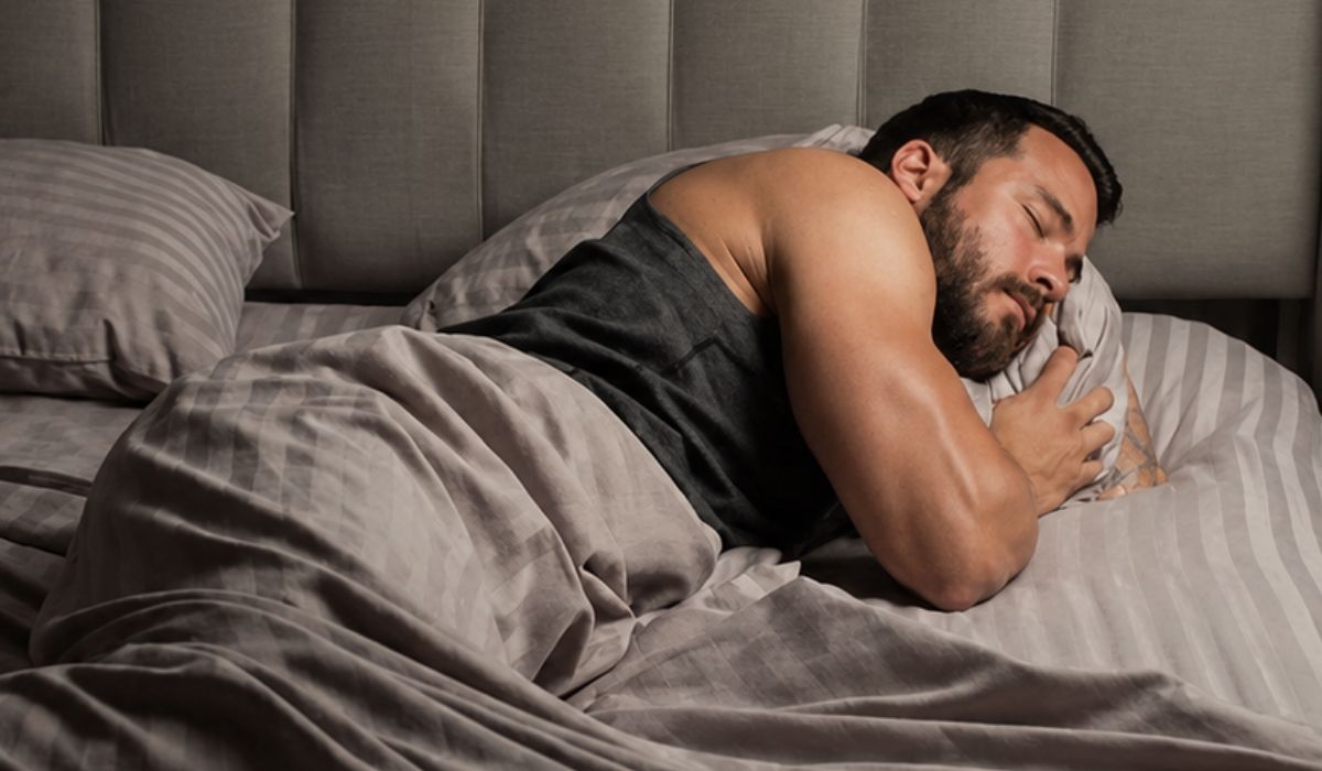 Homem musculoso dormindo de lado em cama confortável, ilustrando a relação entre sono anabólico, hipertrofia e recuperação muscular