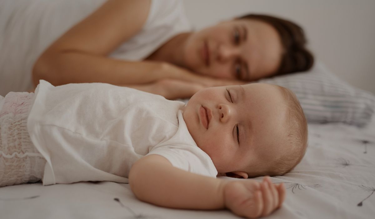 Bebê dormindo tranquilamente na cama com mãe ao fundo, ilustrando curiosidades sobre o sono do bebê e sua rotina de descanso