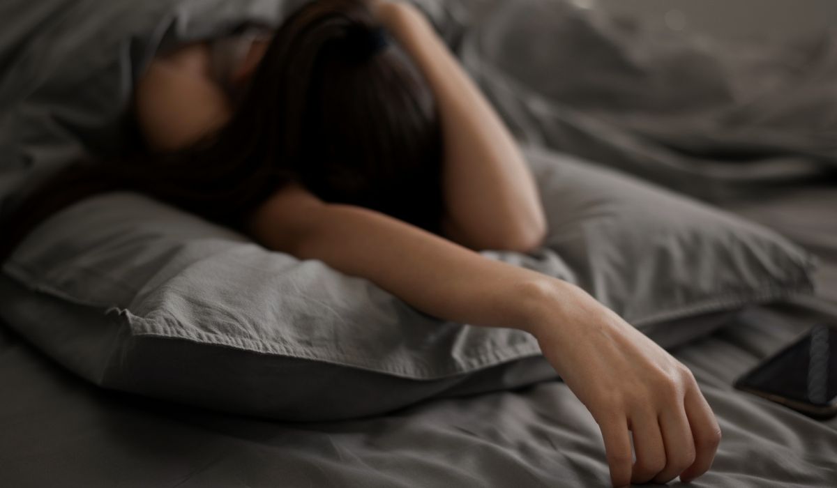 Pessoa deitada na cama com aparência cansada e celular ao lado, ilustrando o tema dormir muito faz mal e os impactos do excesso de sono