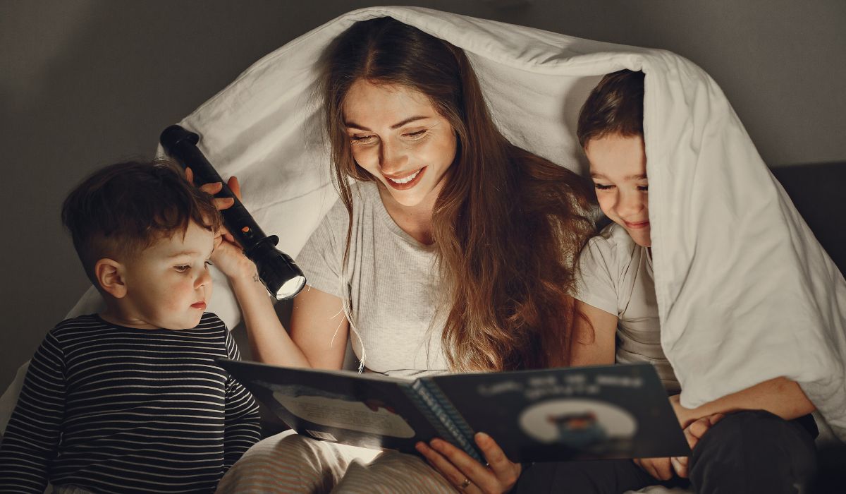 Mãe lendo história infantil para dormir com dois filhos embaixo do cobertor, criando uma rotina noturna acolhedora