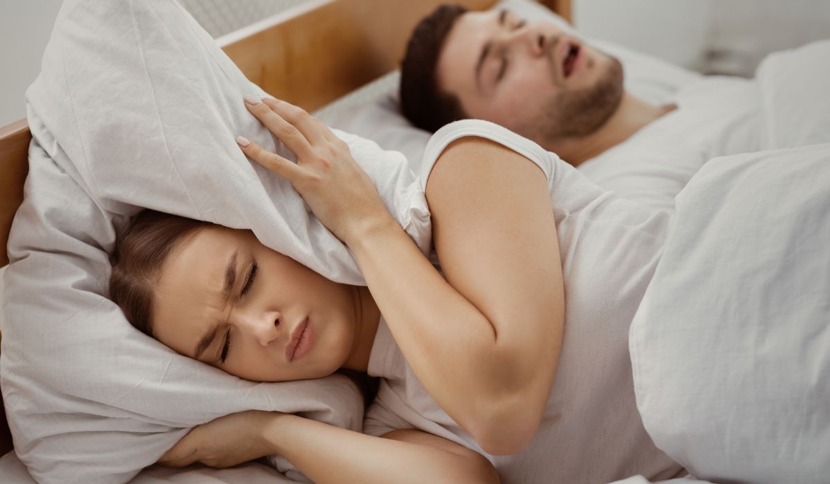 Mulher cobrindo o ouvido com travesseiro ao lado de homem roncando na cama, ilustrando ronco, causas e soluções para melhorar o sono