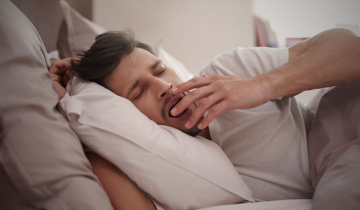 Homem bocejando ao acordar na cama, ilustrando por que dormimos biologia e a importância do sono para o corpo