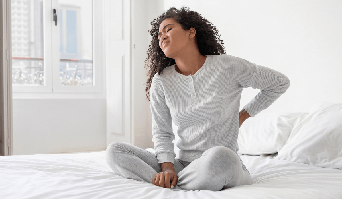 Mulher sentada na cama com a mão na lombar, ilustrando dor nas costas ao acordar e suas possíveis causas