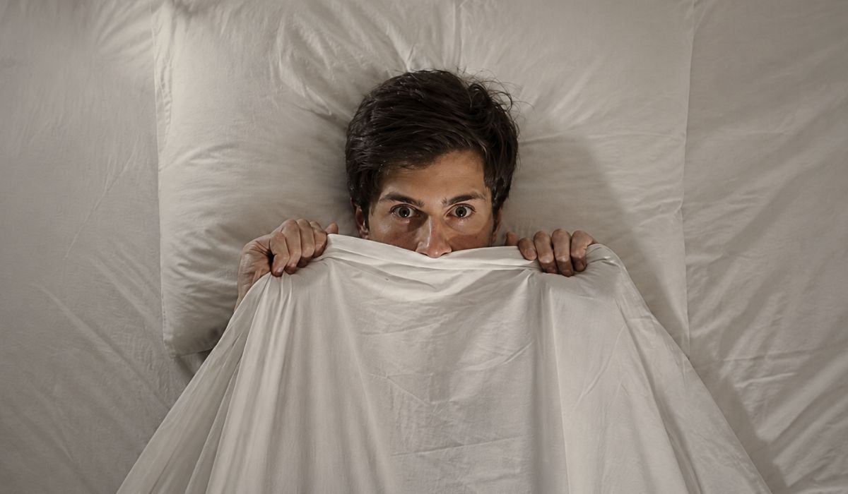 Homem assustado na cama segurando o lençol, ilustrando susto ao dormir e o que é essa sensação durante o sono