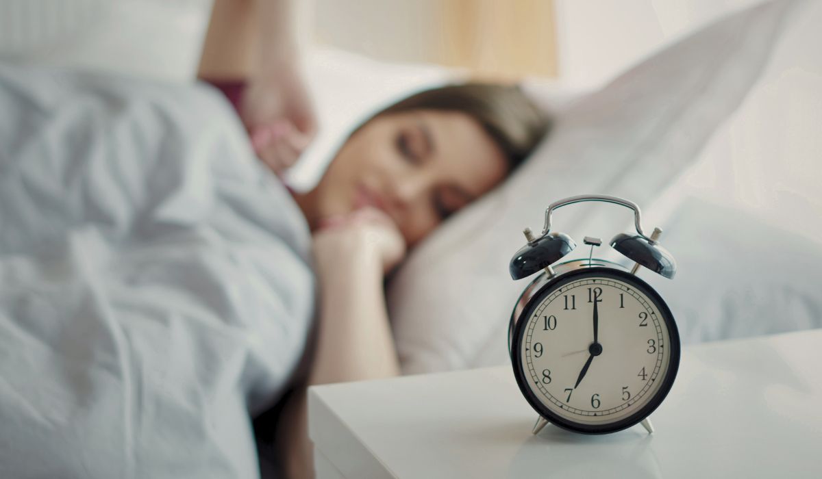 Mulher dormindo com despertador ao lado da cama, ilustrando dicas de como regular o sono e criar uma rotina de descanso saudável.