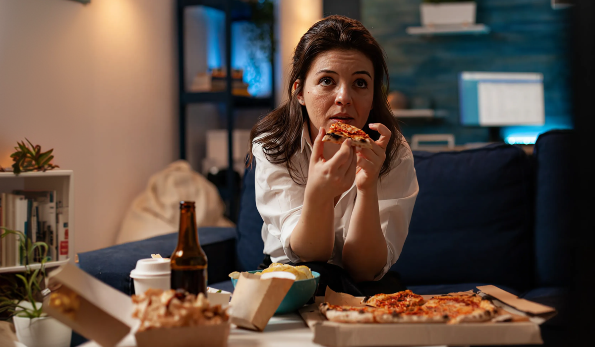Mulher comendo pizza à noite em frente à televisão, ilustrando alimentos que tiram o sono e hábitos noturnos que prejudicam o descanso