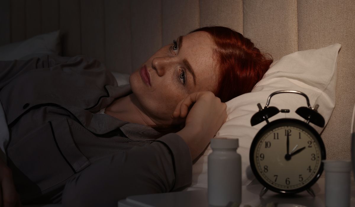 Mulher deitada na cama, acordada durante a noite, ao lado de um relógio e frascos sobre a mesa, ilustrando o tema sonambulismo.