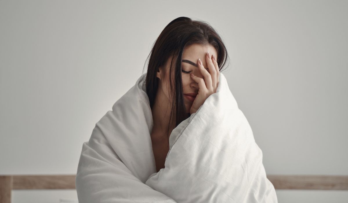 Mulher sentada na cama envolta em cobertor branco, com expressão de cansaço, ilustrando causas de acordar cansado sempre.