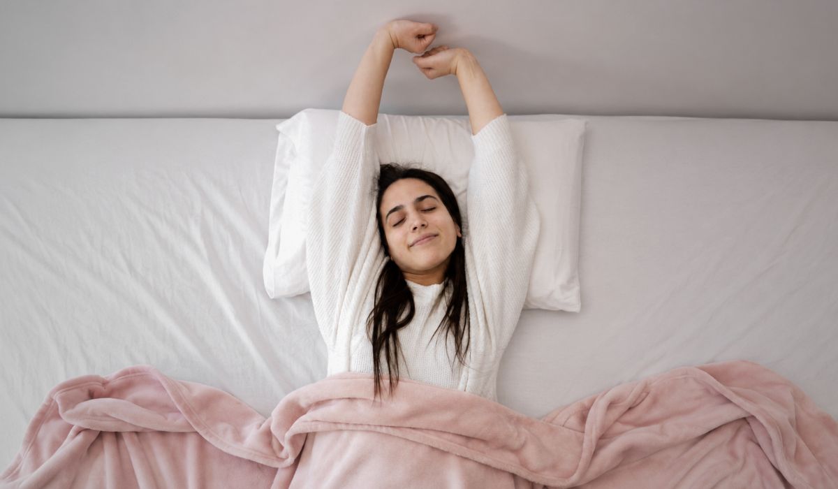Mulher acordando bem-disposta e se espreguiçando na cama, ilustrando a importância de dormir bem para a saúde e o bem-estar.
