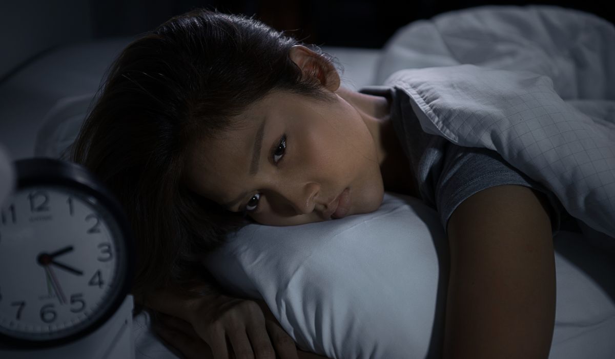 Mulher deitada na cama durante a noite, com olhar cansado e desperto ao lado de um relógio, representando distúrbios do sono, tipos e sintomas.