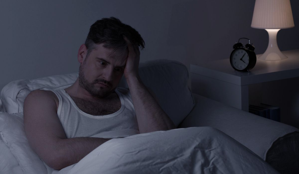 Homem sentado na cama durante a noite, com expressão de cansaço e preocupação, em um quarto escuro com abajur e relógio ao lado.