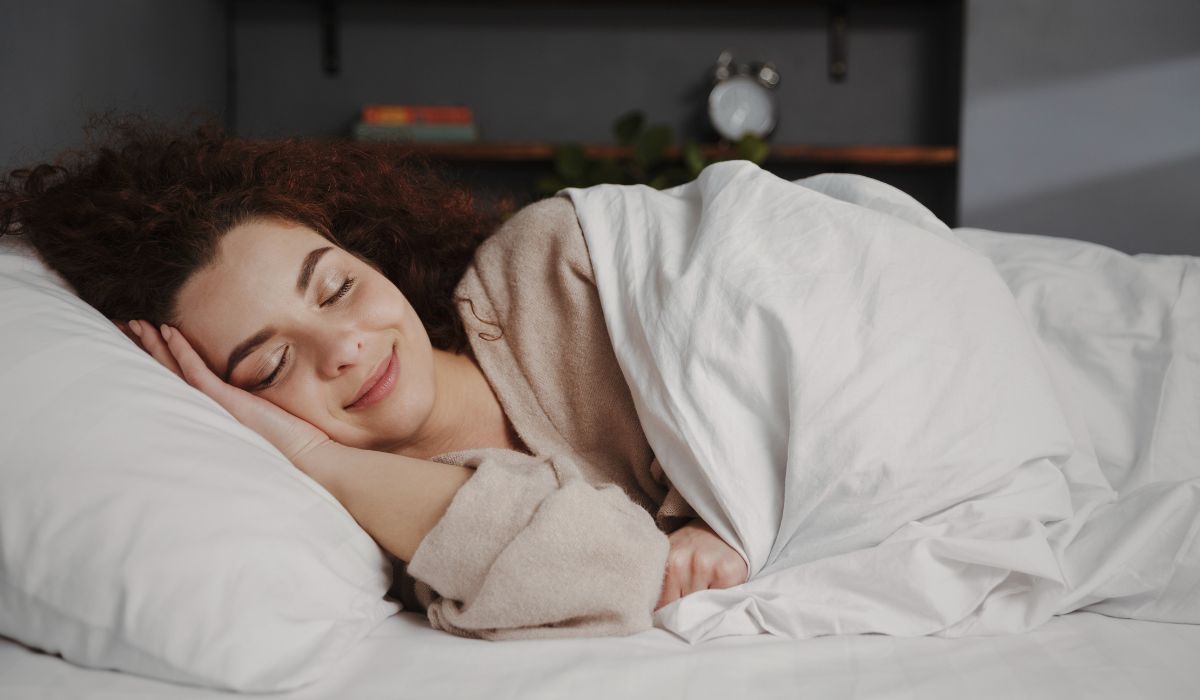 Mulher dormindo tranquilamente em uma cama confortável, com a cabeça apoiada em um travesseiro branco e coberta por um edredom.