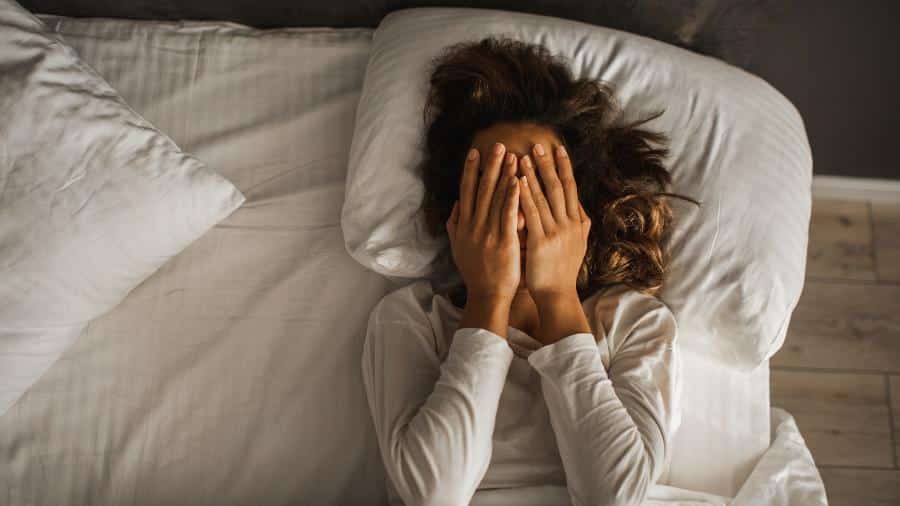 Mulher deitada na cama com as mãos cobrindo o rosto, transmitindo angústia e dificuldade para dormir, representando a paralisia do sono.