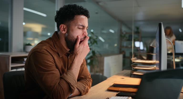 Homem bocejando em frente ao computador no ambiente de trabalho, demonstrando cansaço e sonolência durante o expediente