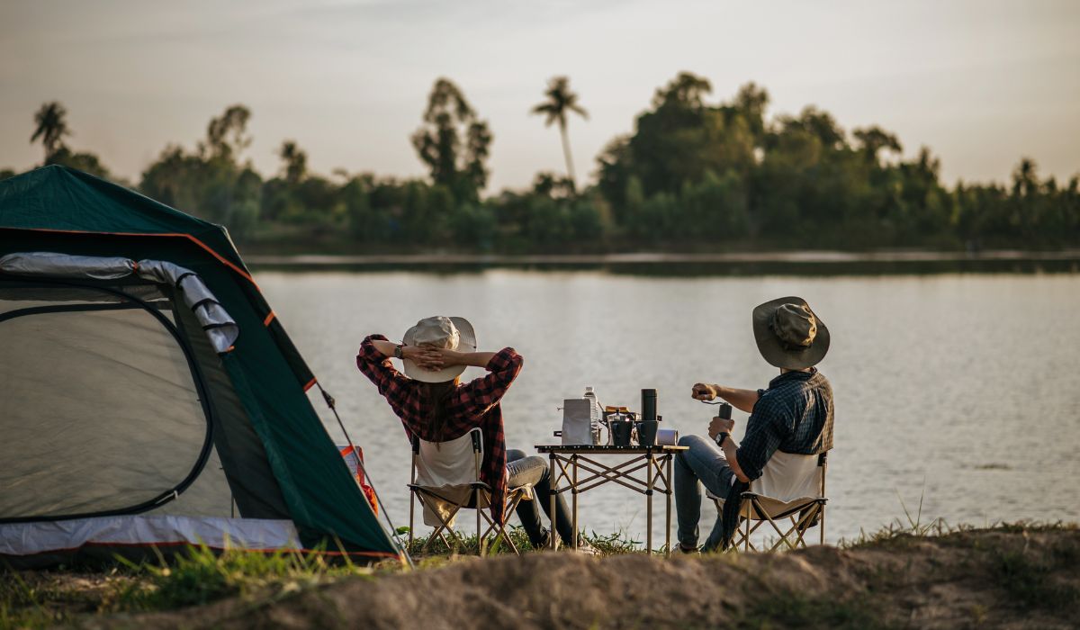 Casal relaxando em cadeiras ao lado de uma barraca, em frente a um lago, durante um acampamento ao ar livre.