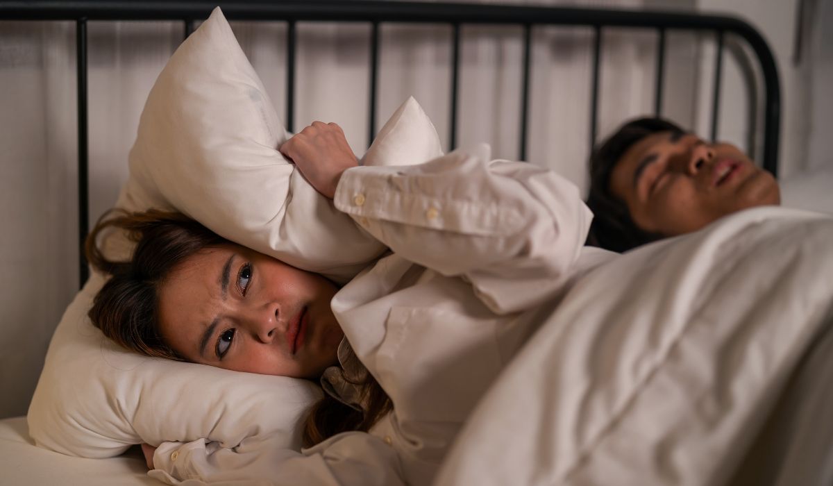 Mulher deitada na cama cobrindo o ouvido com um travesseiro enquanto o parceiro dorme e ronca ao lado.