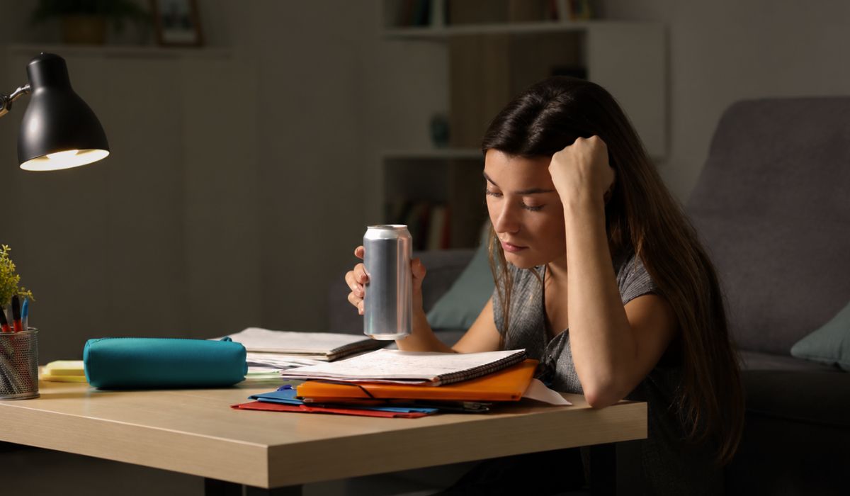 Mulher estudando à noite, com aparência cansada, segurando uma lata de energético enquanto lê anotações na mesa.