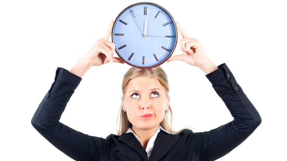 Mulher segurando um relógio redondo acima da cabeça enquanto olha para cima com expressão de dúvida, representando a relação entre tempo e ritmo biológico.