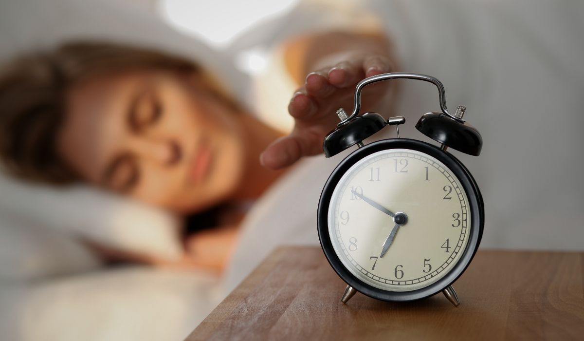 Mulher tentando desligar o despertador ao acordar, ilustrando cronotipo, ciclo circadiano e como o relógio biológico influencia o sono.