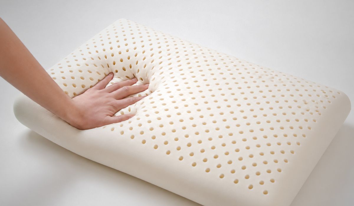 Mão pressionando travesseiro de látex perfurado, ilustrando conforto, elasticidade e ventilação entre as vantagens dos travesseiros de látex.