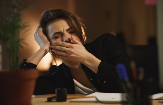 Mulher cansada e com sono bocejando enquanto trabalha à noite, demonstrando dificuldade para dormir e sinais de fadiga.