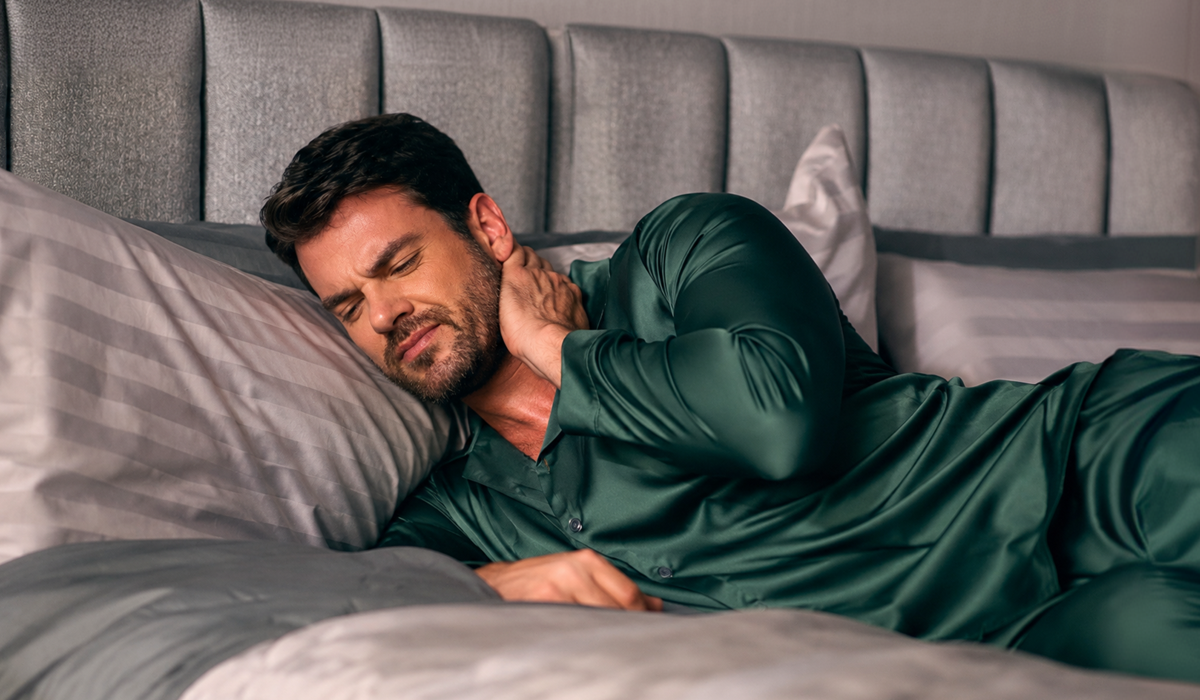 Homem deitado de lado na cama segurando o pescoço com expressão de desconforto, ilustrando o problema de acordar com dor no pescoço.