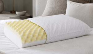 Travesseiro massageador com espuma em gomos e corte interno aparente, apoiado sobre a cama em ambiente neutro e aconchegante.