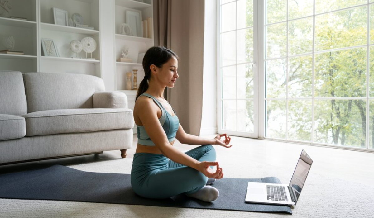 Mulher praticando meditação guiada em casa, sentada sobre um tapete de yoga, com os olhos fechados e um notebook à frente, em um ambiente tranquilo e iluminado